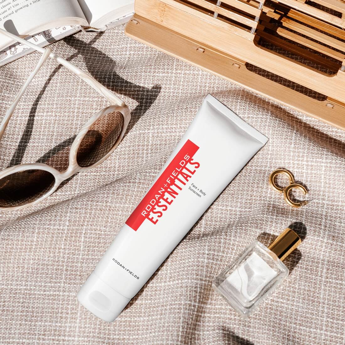 Rodan + Fields Essentials Sunscreen on a beach blanket alongside sunglasses, earrings and other beach items