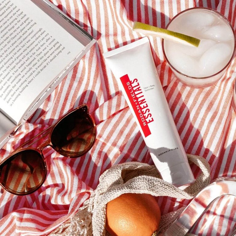 Rodan + Fields Essentials Face & Body Sunscreen on a beach blanket along with sunglasses, cocktail and book