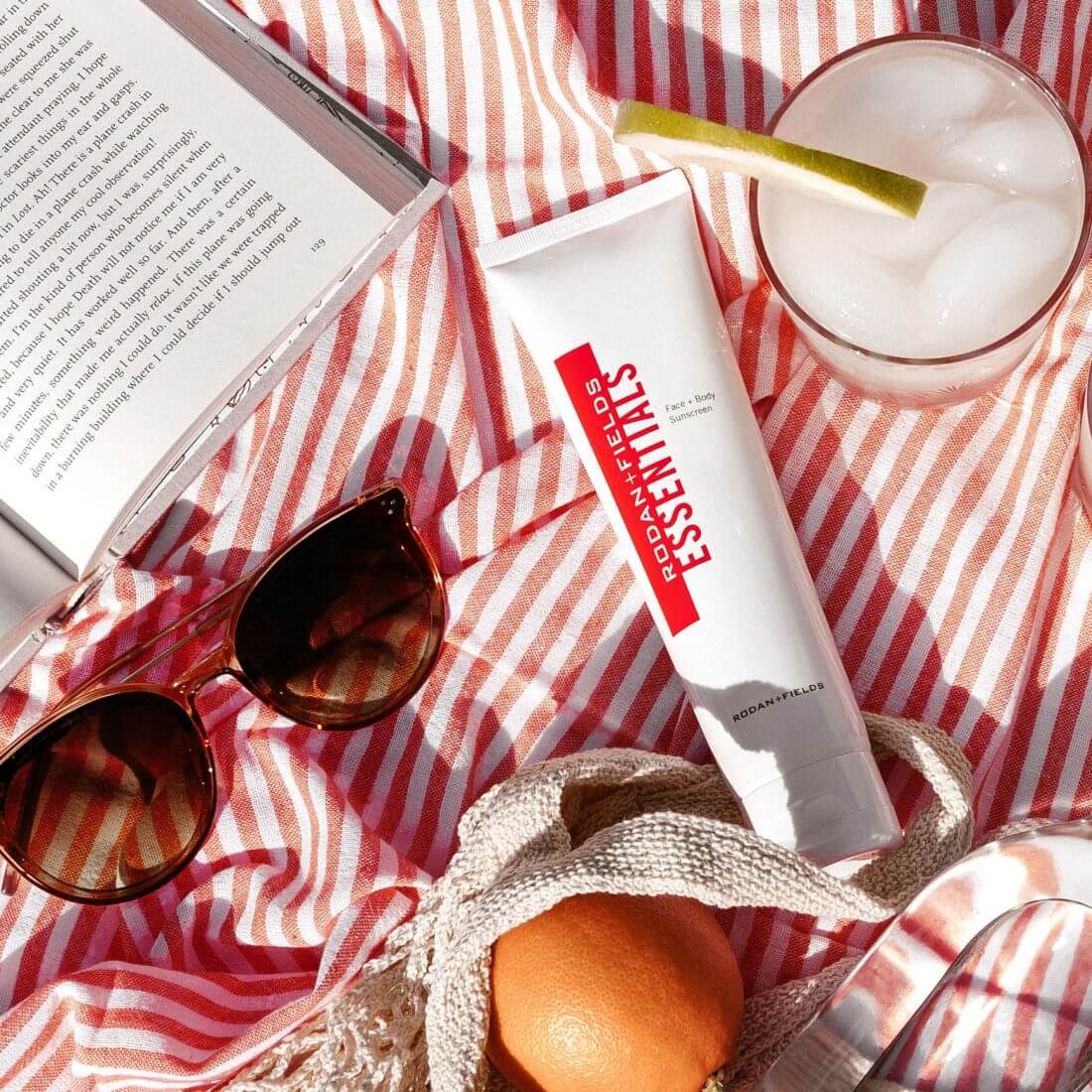 Rodan + Fields Essentials Face & Body Sunscreen on a beach blanket along with sunglasses, cocktail and book