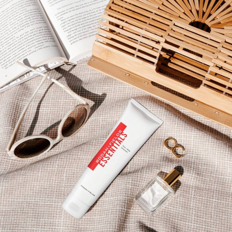 Rodan + Fields Essentials Sunscreen on a beach blanket alongside sunglasses, earrings and other beach items