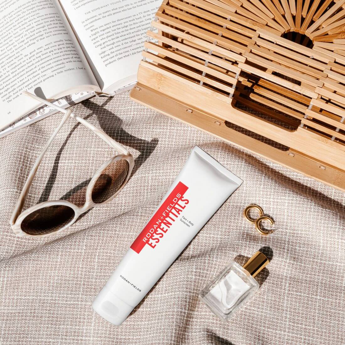 Rodan + Fields Essentials Sunscreen on a beach blanket alongside sunglasses, earrings and other beach items