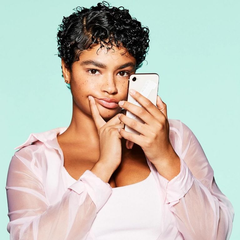 Young woman with short curly hair and dark skin holding up her smartphone with a hand to her face