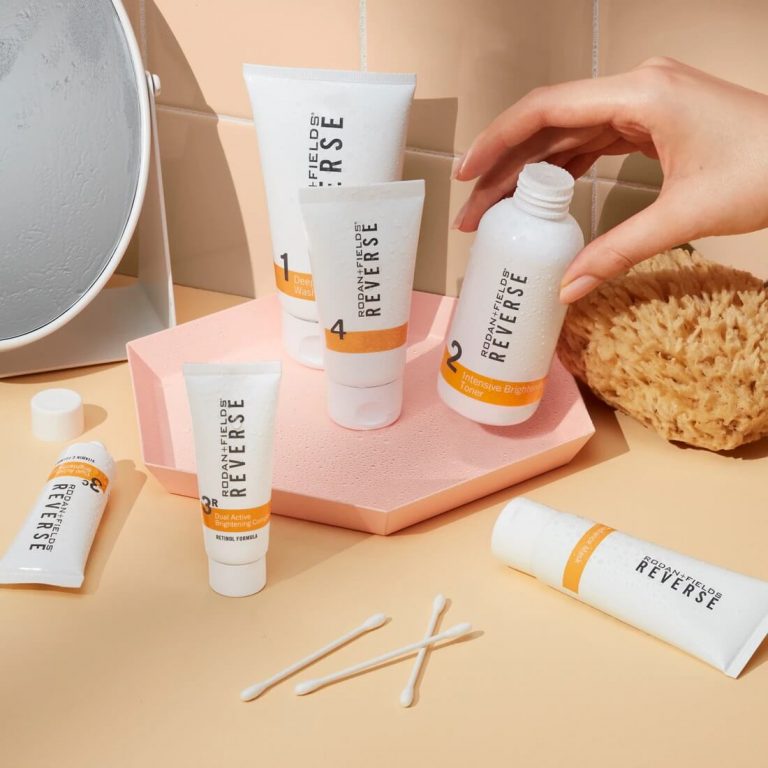 A woman's hand reaching for a bottle of Rodan + Fields Reverse Intensive Brightening toner, with the full Reverse Regimen beside