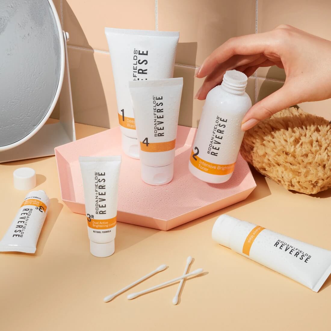 A woman's hand reaching for a bottle of Rodan + Fields Reverse Intensive Brightening toner, with the full Reverse Regimen beside