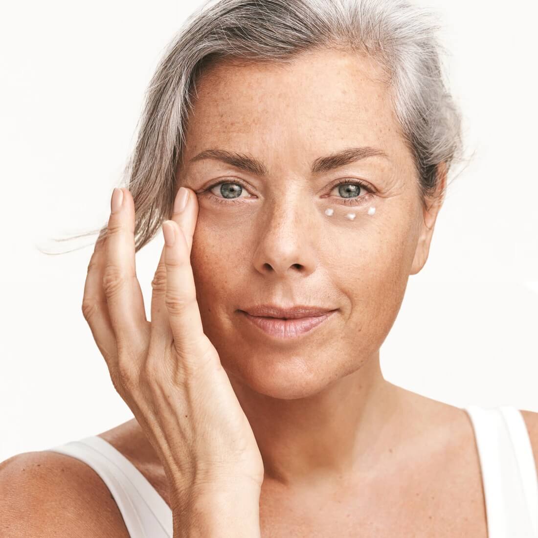 Grey-haired woman gently applying skincare product to her eye area