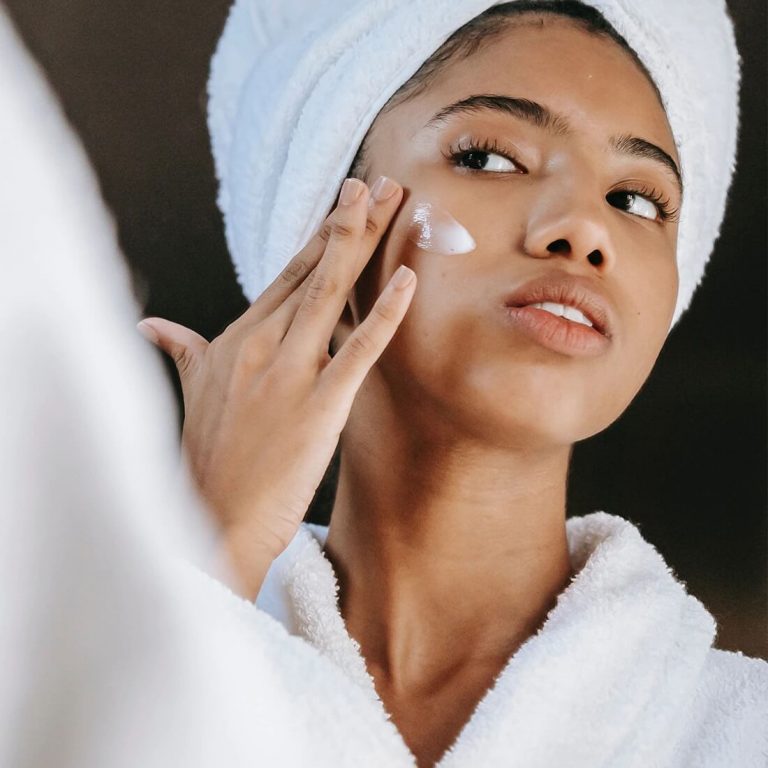 Young woman with hair wrap applying toner to her face in the mirror