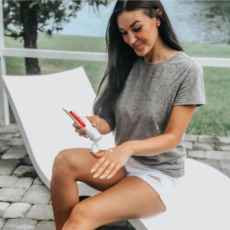 Young woman with dark hair applying Rodan + Fields Essentials SPF to the back of her hand, seated on an outdoor lounge chair