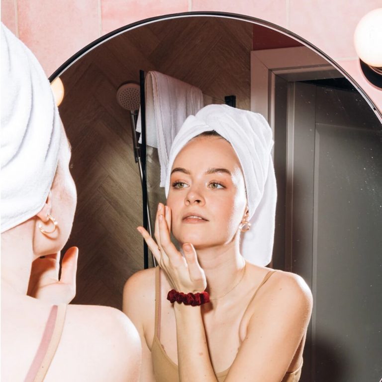 Young woman with hair wrap applying product to her face in a large bathroom mirror