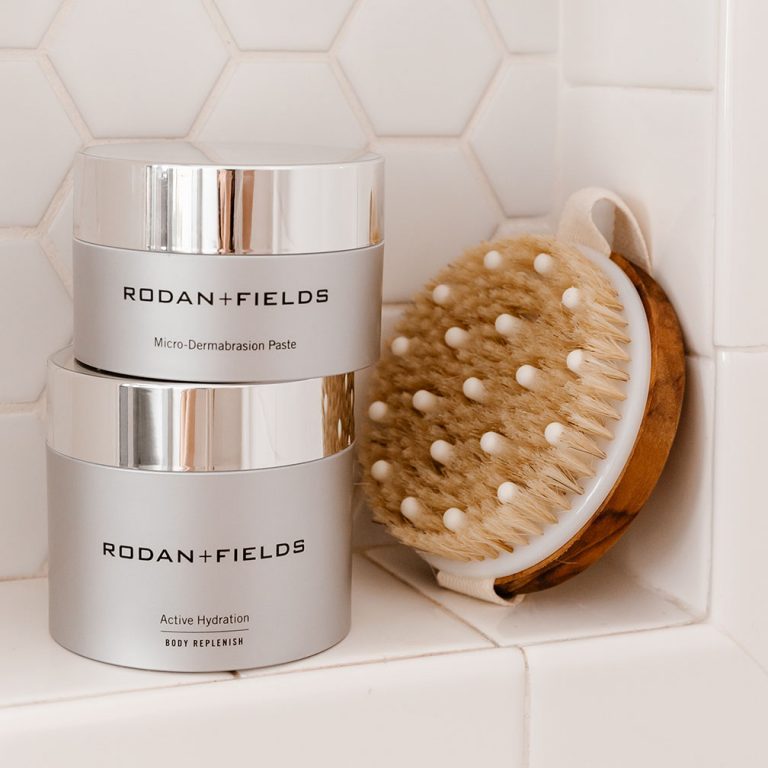 Rodan + Fields Microdermabrasion Paste and Active Hydration Body Replenish products on a shower ledge alongside scrubbing brush