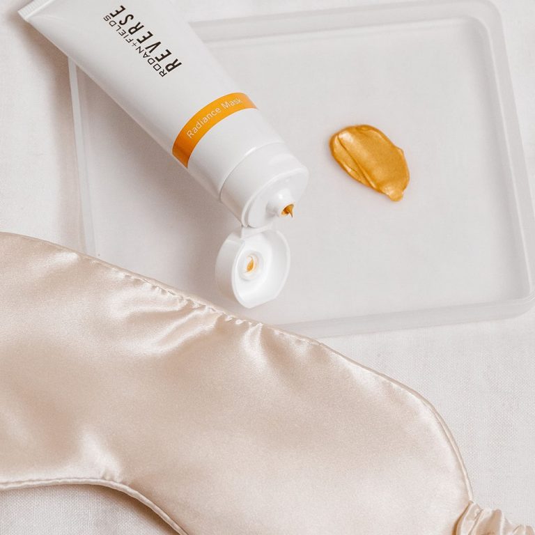 Open tube of Rodan + Fields Reverse Radiance Mask showing golden product color and texture, alongside a sleeping eye mask.