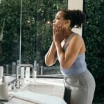 Woman in a bathroom looking in the mirror while applying R+F skincare products.
