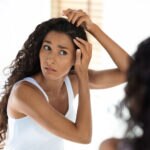 Young woman with dark wavy hair inspects her hair and scalp in the mirror