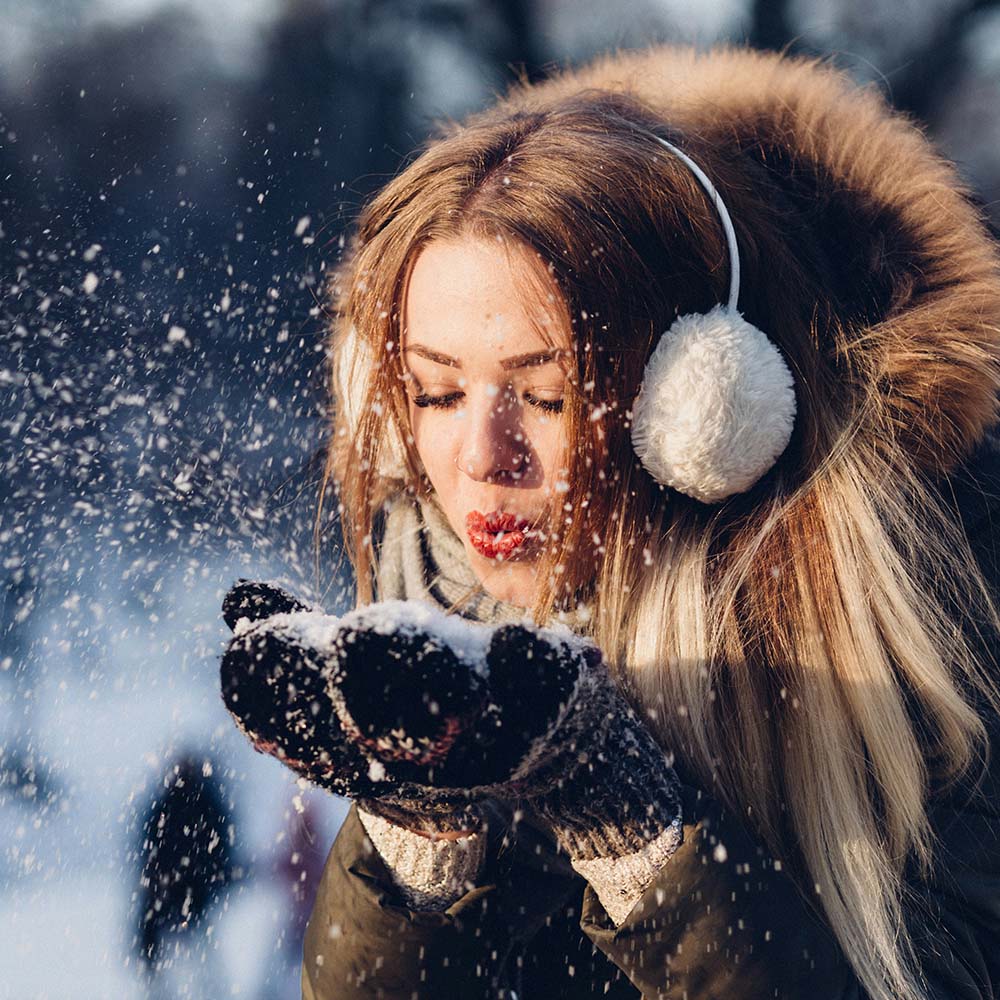 Young woman in an outdoor bright snowy setting with gloves and earmuffs blowing powdery snow from her hands. Photo by freestocks