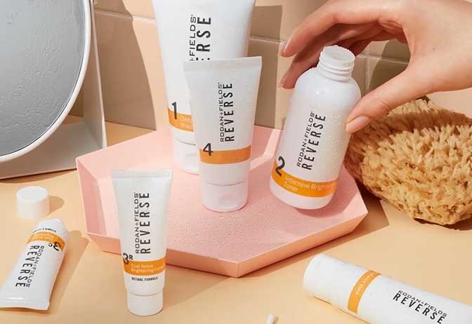 A woman's hand reaching for a bottle of Rodan + Fields Reverse Intensive Brightening toner, with the full Reverse Regimen beside. A woman's hand reaching for a bottle of Rodan + Fields Reverse Intensive Brightening toner, with the full Reverse Regimen beside.